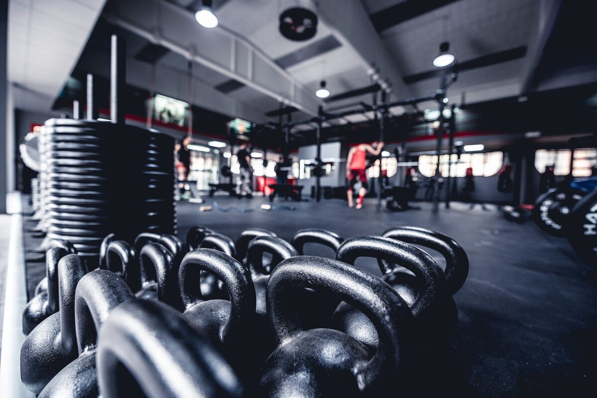 Kettlebell neri in fila e dischi impilati in primo piano, rack con atleti sullo sfondo in una sala con luci a sospensione