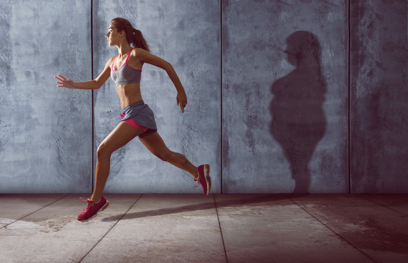 Ragazza che corre in top grigio e pantaloncini rosa davanti a un muro di cemento con l'ombra di una figura più robusta
